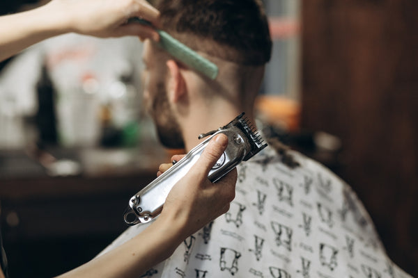 Hairdresser_Cutting_Bearded_Young_Man's_Hair_With_Trimmer_and_Combing_Hair_On_Head